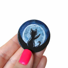 Load image into Gallery viewer, A lightweight wooden disc features art of the silhouette of a black cat, with a front paw reaching up, sitting in front of a big glowing full moon. Available as a keepsake, magnet or necklace
