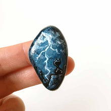 Load image into Gallery viewer, a beach stone with cat art: a painting of a black cat on a cliff, looking out into the night sky, filled with lightning. this piece is available as a keepsake, magnet or a pendant necklace
