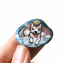 Load image into Gallery viewer, a small smooth beach stone, hand painted with the portrait of an akita dog, painted as an angel, it peers out over fluffy clouds against a blue and pink sky. this piece is available as a keepsake or pendant necklace
