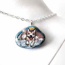 Load image into Gallery viewer, a small smooth beach stone, hand painted with the portrait of an akita dog, painted as an angel, it peers out over fluffy clouds against a blue and pink sky. this piece is available as a keepsake or pendant necklace
