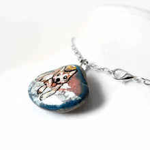 Load image into Gallery viewer, a small smooth beach stone, hand painted with the portrait of an akita dog, painted as an angel, it peers out over fluffy clouds against a blue and pink sky. this piece is available as a keepsake or pendant necklace
