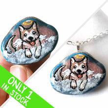 Load image into Gallery viewer, a small smooth beach stone, hand painted with the portrait of an akita dog, painted as an angel, it peers out over fluffy clouds against a blue and pink sky. this piece is available as a keepsake or pendant necklace
