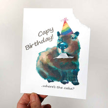 Load image into Gallery viewer, a greeting card featuring an illustration of a capybara wearing a rainbow birthday hat. The top right corner of the card hangs from its mouth. the front reads, Capy Birthday! ..where's the cake?
