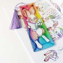 Load image into Gallery viewer, A PET (polyethylene terephthalate) bookmark and purple tassel with a small bead, featuring a character from Rainbow of Crazy Comics reading in a cozy, colourful box: an English spot rabbit reads a book with a ghost holding a camera on the cover. on the side, it says @ Rainbowofcrazy.
