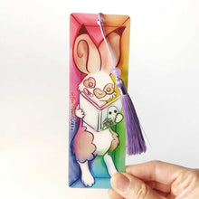 Load image into Gallery viewer, A PET (polyethylene terephthalate) bookmark and purple tassel with a small bead, featuring a character from Rainbow of Crazy Comics reading in a cozy, colourful box: an English spot rabbit reads a book with a ghost holding a camera on the cover. on the side, it says @ Rainbowofcrazy.
