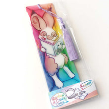 Load image into Gallery viewer, A PET (polyethylene terephthalate) bookmark and purple tassel with a small bead, featuring a character from Rainbow of Crazy Comics reading in a cozy, colourful box: an English spot rabbit reads a book with a ghost holding a camera on the cover. on the side, it says @ Rainbowofcrazy. The bookmark is packaged in a sleeve with a small band with the text "Rainbow of Crazy Comics bookmark"
