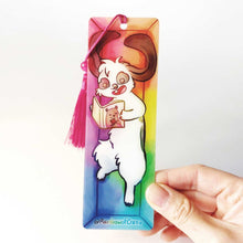 Load image into Gallery viewer, A PET (polyethylene terephthalate) bookmark and red tassel with a small bead, featuring a character from Rainbow of Crazy Comics reading in a cozy, colourful box: a mini lop rabbit reads a book bear holding a hat on the cover. on the bottom, it says @ Rainbowofcrazy.
