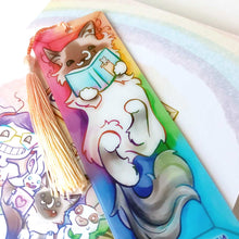 Load image into Gallery viewer, A PET (polyethylene terephthalate) bookmark and orange/yellow tassel, featuring a character from Rainbow of Crazy Comics: a ragdoll cat reads a book with a cat on the cover, as she lounges in a colourful box. on the bottom, it says @ Rainbowofcrazy
