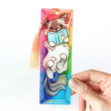 Load image into Gallery viewer, A PET (polyethylene terephthalate) bookmark and orange/yellow tassel, featuring a character from Rainbow of Crazy Comics: a ragdoll cat reads a book with a cat on the cover, as she lounges in a colourful box. on the bottom, it says @ Rainbowofcrazy
