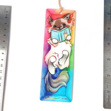 Load image into Gallery viewer, A PET (polyethylene terephthalate) bookmark and orange/yellow tassel, featuring a character from Rainbow of Crazy Comics: a ragdoll cat reads a book with a cat on the cover, as she lounges in a colourful box. on the bottom, it says @ Rainbowofcrazy
