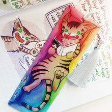 Load image into Gallery viewer, A PET (polyethylene terephthalate) bookmark and white tassel, featuring a character from Rainbow of Crazy Comics reading in a cozy, colourful box: a tabby cat reads a book with a cat on the cover. on the bottom, it says @ Rainbowofcrazy
