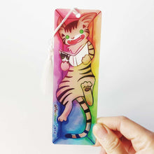 Load image into Gallery viewer, A PET (polyethylene terephthalate) bookmark and white tassel, featuring a character from Rainbow of Crazy Comics reading in a cozy, colourful box: a tabby cat reads a book with a cat on the cover. on the bottom, it says @ Rainbowofcrazy
