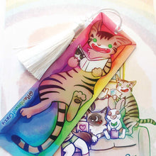 Load image into Gallery viewer, A PET (polyethylene terephthalate) bookmark and white tassel, featuring a character from Rainbow of Crazy Comics reading in a cozy, colourful box: a tabby cat reads a book with a cat on the cover. on the bottom, it says @ Rainbowofcrazy
