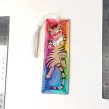 Load image into Gallery viewer, A PET (polyethylene terephthalate) bookmark and white tassel, featuring a character from Rainbow of Crazy Comics reading in a cozy, colourful box: a tabby cat reads a book with a cat on the cover. on the bottom, it says @ Rainbowofcrazy.
