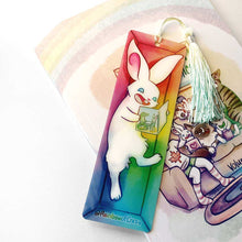 Load image into Gallery viewer, A PET (polyethylene terephthalate) bookmark and yellow/green tassel, featuring a character from Rainbow of Crazy Comics: a white rabbit reads a book with a crocodile and rabbit on the cover, as she lounges in a colourful box. on the bottom, it says @ Rainbowofcrazy
