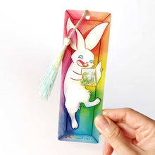 Load image into Gallery viewer, A PET (polyethylene terephthalate) bookmark and yellow/green tassel, featuring a character from Rainbow of Crazy Comics: a white rabbit reads a book with a crocodile and rabbit on the cover, as she lounges in a colourful box. on the bottom, it says @ Rainbowofcrazy
