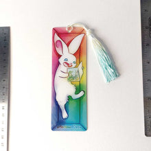 Load image into Gallery viewer, A PET (polyethylene terephthalate) bookmark and yellow/green tassel, featuring a character from Rainbow of Crazy Comics: a white rabbit reads a book with a crocodile and rabbit on the cover, as she lounges in a colourful box. on the bottom, it says @ Rainbowofcrazy
