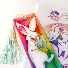 Load image into Gallery viewer, A PET (polyethylene terephthalate) bookmark and yellow/green tassel, featuring a character from Rainbow of Crazy Comics: a white rabbit reads a book with a crocodile and rabbit on the cover, as she lounges in a colourful box. on the bottom, it says @ Rainbowofcrazy
