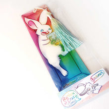 Load image into Gallery viewer, A PET (polyethylene terephthalate) bookmark and yellow/green tassel, featuring a character from Rainbow of Crazy Comics: a white rabbit reads a book with a crocodile and rabbit on the cover, as she lounges in a colourful box. on the bottom, it says @ Rainbowofcrazy. The bookmark is packaged in a sleeve with a small band with the text "Rainbow of Crazy Comics bookmark"
