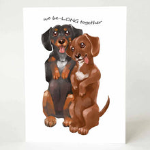 Load image into Gallery viewer, a greeting card with an illustration of one black dachshund, and one brown dachshund, both standing up on their hind legs. it reads, "we be-LONG together"
