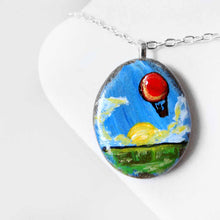Load image into Gallery viewer, a small rock art of a red hot air balloon, floatiing in the sky over a green landscape, sun shining. available as a keepsake or pendant necklace, or magnet
