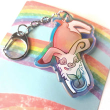 Load image into Gallery viewer, A clear, blue-tinted holographic acrylic keychain featuring a character from Rainbow of Crazy Comics: an English spot rabbit, lying down, admires a potted plant. 
