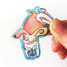Load image into Gallery viewer, A clear, blue-tinted holographic acrylic keychain featuring a character from Rainbow of Crazy Comics: an English spot rabbit, lying down, admires a potted plant. 
