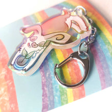 Load image into Gallery viewer, A clear, blue-tinted holographic acrylic keychain featuring a character from Rainbow of Crazy Comics: an English spot rabbit, lying down, admires a potted plant. 
