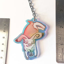 Load image into Gallery viewer, A clear, blue-tinted holographic acrylic keychain featuring a character from Rainbow of Crazy Comics: an English spot rabbit, lying down, admires a potted plant. 2.5" long
