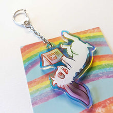 Load image into Gallery viewer, A clear, rainbow holographic acrylic keychain featuring a character from Rainbow of Crazy Comics: a smiling mini lop rabbit lounges on his back as he reads a book with a picture of a rabbit on the back.
