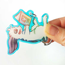 Load image into Gallery viewer, A clear, rainbow holographic acrylic keychain featuring a character from Rainbow of Crazy Comics: a smiling mini lop rabbit lounges on his back as he reads a book with a picture of a rabbit on the back.
