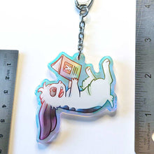 Load image into Gallery viewer, A clear, rainbow holographic acrylic keychain featuring a character from Rainbow of Crazy Comics: a smiling mini lop rabbit lounges on his back as he reads a book with a picture of a rabbit on the back. 2.5" large.
