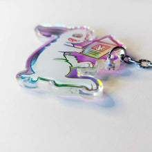 Load image into Gallery viewer, A clear, rainbow holographic acrylic keychain featuring a character from Rainbow of Crazy Comics: a smiling mini lop rabbit lounges on his back as he reads a book with a picture of a rabbit on the back. Shown at a different angle.
