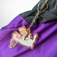 Load image into Gallery viewer, A clear, rainbow holographic acrylic keychain featuring a character from Rainbow of Crazy Comics: a smiling mini lop rabbit lounges on his back as he reads a book with a picture of a rabbit on the back. Hanging from a purple bag.
