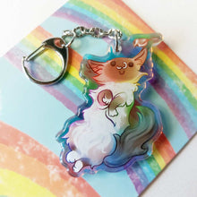 Load image into Gallery viewer, A clear, rainbow holographic acrylic keychain featuring a character from Rainbow of Crazy Comics: a smiling fluffy ragdoll cat, lying on her back, with a tiny black-capped chickadee, the bird perched on the cat's ear.
