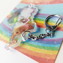 Load image into Gallery viewer, A clear, rainbow holographic acrylic keychain featuring a character from Rainbow of Crazy Comics: a smiling fluffy ragdoll cat, lying on her back, with a tiny black-capped chickadee, the bird perched on the cat's ear.
