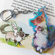Load image into Gallery viewer, A clear, rainbow holographic acrylic keychain featuring a character from Rainbow of Crazy Comics: a smiling fluffy ragdoll cat, lying on her back, with a tiny black-capped chickadee, the bird perched on the cat's ear. The keychain is placed next to an illustration of the same cat, with a white rabbit on the cat's back.
