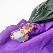Load image into Gallery viewer, A clear, rainbow holographic acrylic keychain featuring a character from Rainbow of Crazy Comics: a smiling fluffy ragdoll cat, lying on her back, with a tiny black-capped chickadee, the bird perched on the cat's ear. It hangs from a purple backpack.
