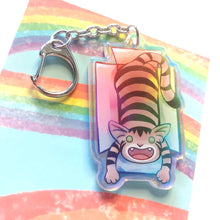 Load image into Gallery viewer, A clear, blue tinted holographic acrylic keychain featuring a character from Rainbow of Crazy Comics: a smiling tabby cat lounges in a cardboard box.
