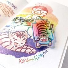 Load image into Gallery viewer, A clear, blue tinted holographic acrylic keychain featuring a character from Rainbow of Crazy Comics: a smiling tabby cat lounges in a cardboard box.
