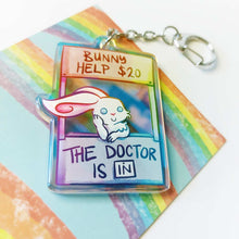 Load image into Gallery viewer, A clear, rainbow holographic acrylic keychain featuring a character from Rainbow of a Crazy Comics: a smiling white rabbit at a homemade booth that says "bunny help $20" at the top, and "the doctor is in" at the bottom. 
