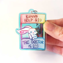 Load image into Gallery viewer, A clear, rainbow holographic acrylic keychain featuring a character from Rainbow of a Crazy Comics: a smiling white rabbit at a homemade booth that says "bunny help $20" at the top, and "the doctor is in" at the bottom. 
