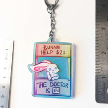 Load image into Gallery viewer, A clear, rainbow holographic acrylic keychain featuring a character from Rainbow of a Crazy Comics: a smiling white rabbit at a homemade booth that says "bunny help $20" at the top, and "the doctor is in" at the bottom. 2.5"
