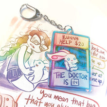 Load image into Gallery viewer, A clear, rainbow holographic acrylic keychain featuring a character from Rainbow of a Crazy Comics: a smiling white rabbit at a homemade booth that says "bunny help $20" at the top, and "the doctor is in" at the bottom. 
