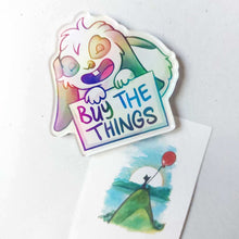 Load image into Gallery viewer, An acrylic magnet featuring a character from Rainbow of Crazy Comics: a mini lop rabbit smiles and holds up a sign that says "BUY THE THINGS". The Magnet holds up a small print of a black cat sitting on top of a hill, holding a red balloon.
