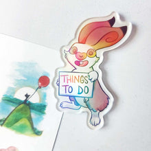 Load image into Gallery viewer, An acrylic magnet featuring a character from Rainbow of Crazy Comics: a rabbit smiles and holds up a sign that says "THINGS TO DO". The Magnet holds up a small print of a black cat sitting on top of a hill, holding a red balloon.
