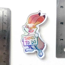 Load image into Gallery viewer, An acrylic magnet featuring a character from Rainbow of Crazy Comics: a rabbit smiles and holds up a sign that says "THINGS TO DO". The Magnet holds up a small print of a black cat sitting on top of a hill, holding a red balloon.
