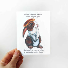 Load image into Gallery viewer, a greeting card, featuring an illustration of a brown and white rabbit with a pancake on its head. front reads, "I didn't know which card to get you / So here's a bunny with a pancake on its head"
