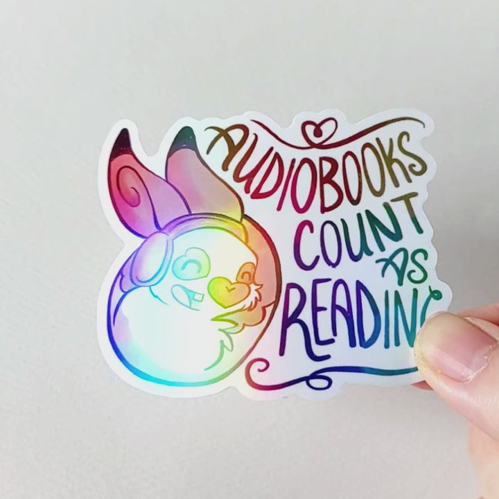 A vinyl rainbow holographic sticker featuring a character from Rainbow of Crazy Comics: a smiling rabbit wearing headphones. the sticker reads, "Audiobooks count as reading"