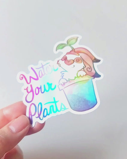 hand tilting a sticker back and forth to show the holographic finish. sticker featuring an english spot rabbit (from Rainbow of Crazy Comics) in a plant pot. a seedling sits on top of the rabbit's head. the sticker has the words "Water Your Plants".